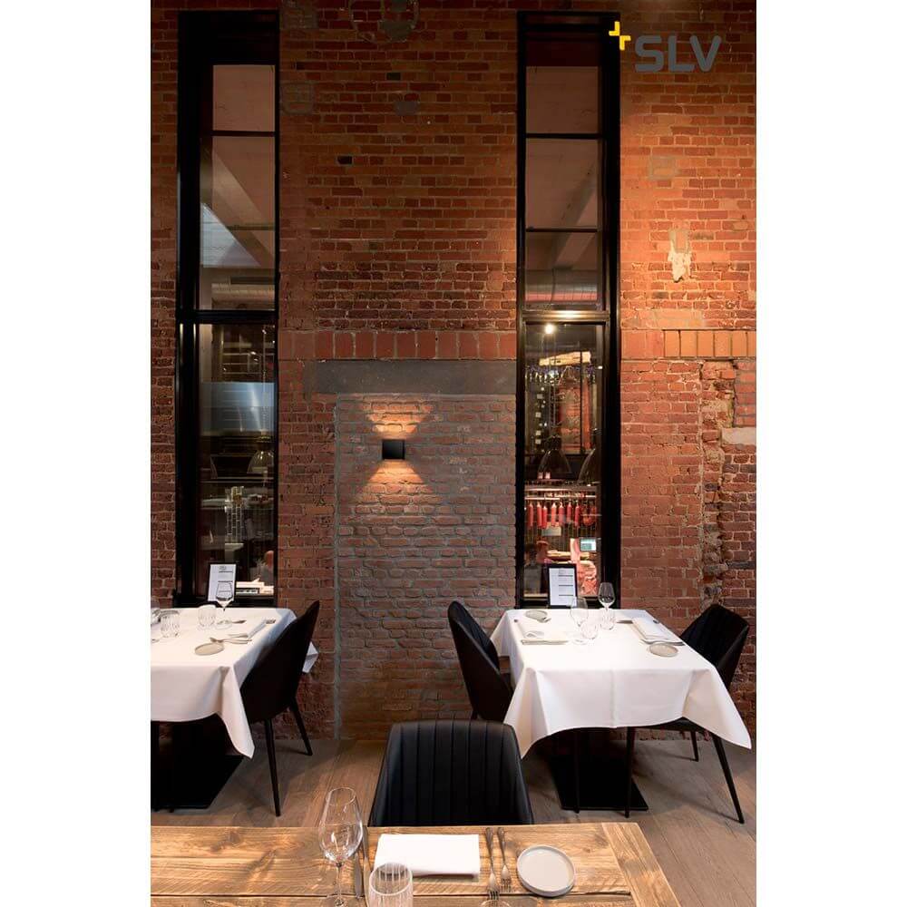 Solid Cube Wandleuchte QT14 Grau beleuchtet einen modernen Restaurantraum mit Ziegelmauer und eleganten Tischen.