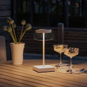 Scilla LED Akku Tischleuchte IP54 auf einem Tisch, umgeben von Pflanzen und Gläsern bei Dämmerung.