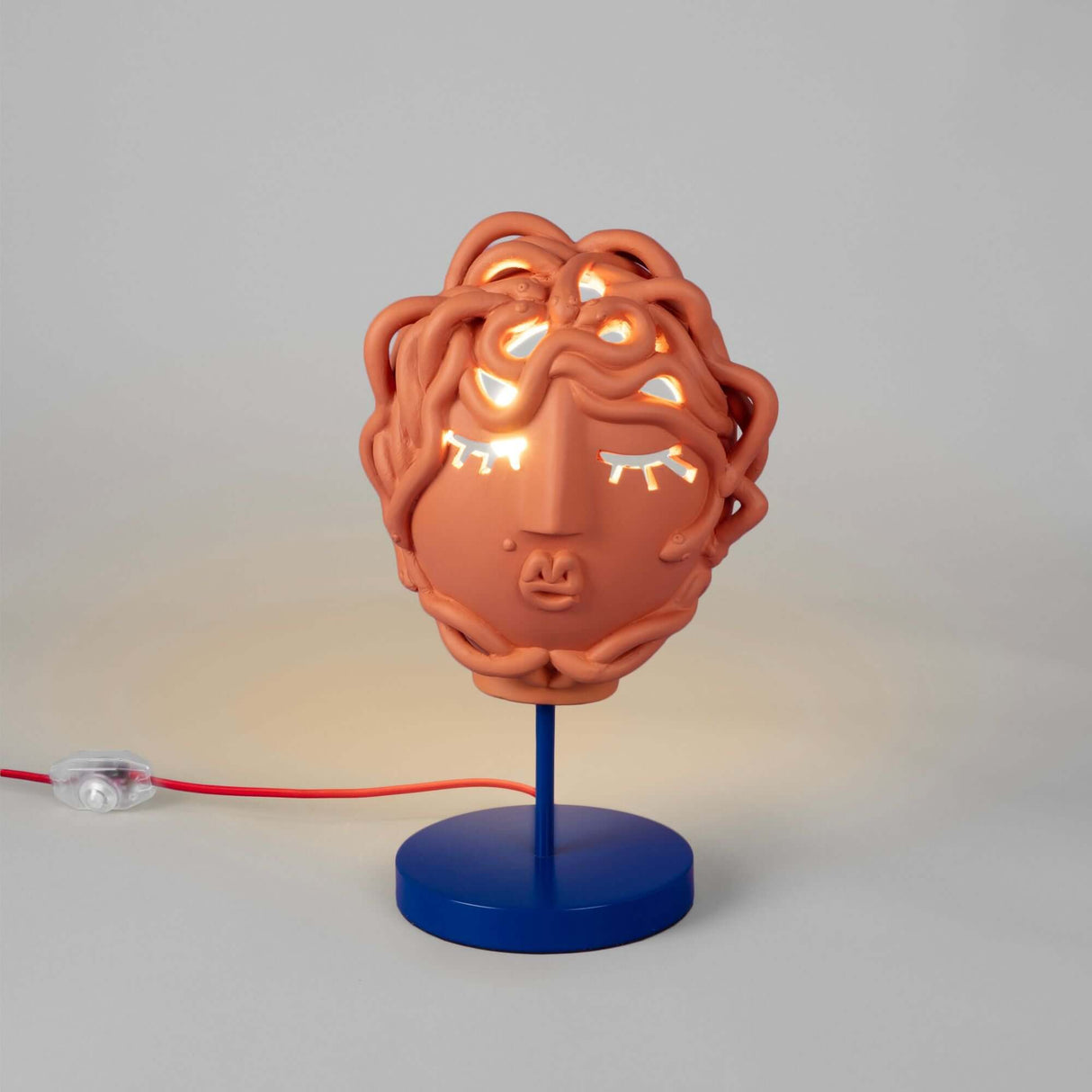 Seletti Magna Graecia Tischleuchte Medusa Maske Terracotta auf blauem Sockel, terrakottafarbene Medusa-Tischlampe mit warmem Licht