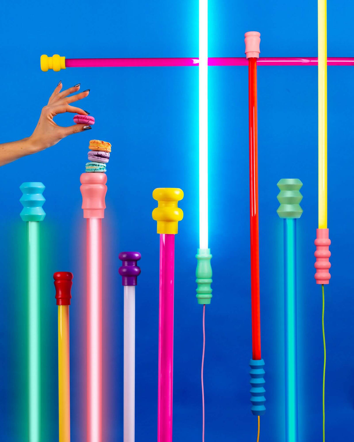 Seletti Superlinea LED-Leuchte 141cm in bunten Neonfarben vor blauem Hintergrund, modernes Design und stapelnde Hand mit Macarons