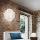 Slamp Wand- & Deckenlampe Veli Foliage an einer Steinwand über einem Bett platziert, mit Fensterblick.