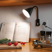 Stikkontaktlampe Junus med fleksarm i svart over et kjøkkenbord med bok, krydder og grønnsaker.