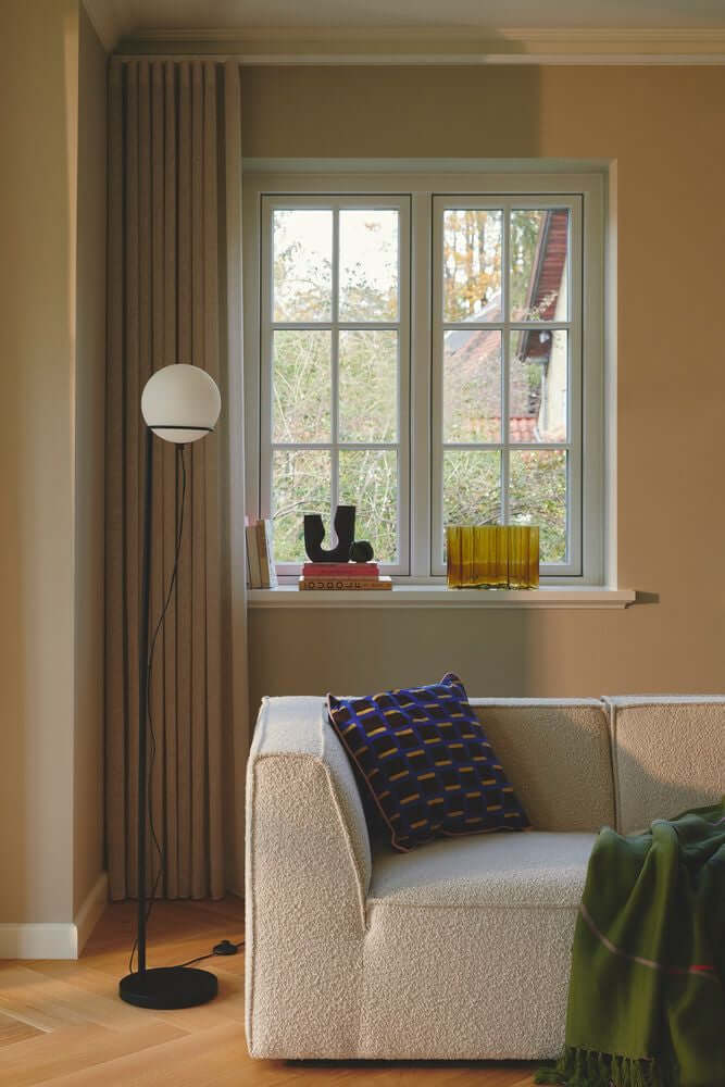 Stehlampe Wilson neben einem Sofa, erhellt den Raum mit sanftem Licht durch den runden Glasschirm.