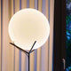 Moderne Stehlampe mit runder, milchiger Glaskugel und elegantem Gestell, vor einem Fenster mit Vorhängen.
