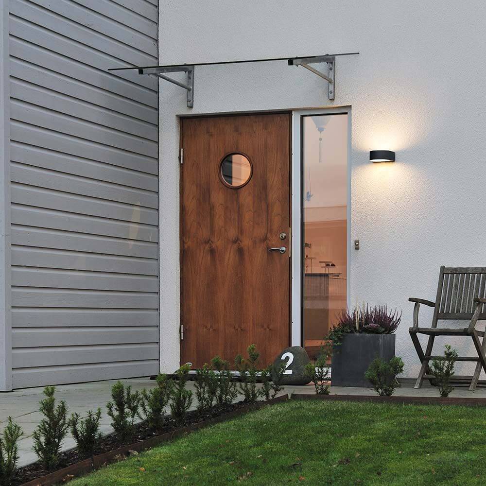 Teramo Außen-Wandleuchte Schwarz, opales Glas neben Eingangstür mit Holz und Garten.
