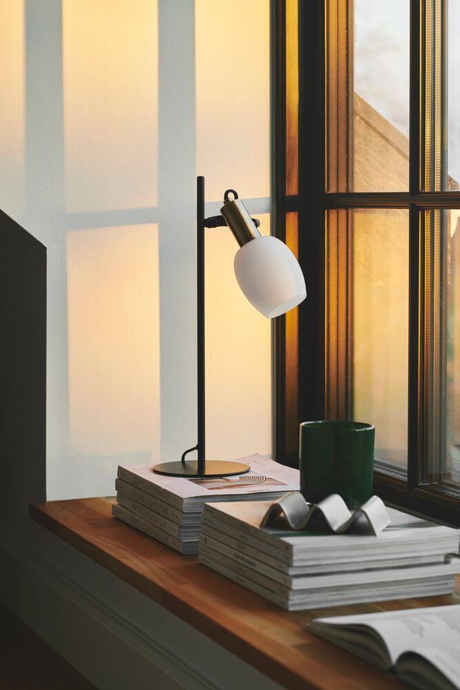 Tischlampe Arild auf einem Holztisch neben mehreren Bookstack und einer grünen Kerze, mit Fenster im Hintergrund.