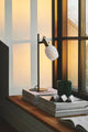 Tischlampe Arild auf einem Holztisch neben mehreren Bookstack und einer grünen Kerze, mit Fenster im Hintergrund.