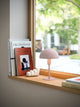 Tischlampe Ellen Mini auf einer Fensterbank mit Büchern und einer dekorativen Figur, skandinavisches Design.