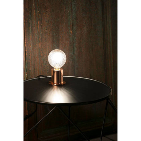 Lampe de table TEN cuivre sur une table noire avec sphère en verre, à la lumière douce.