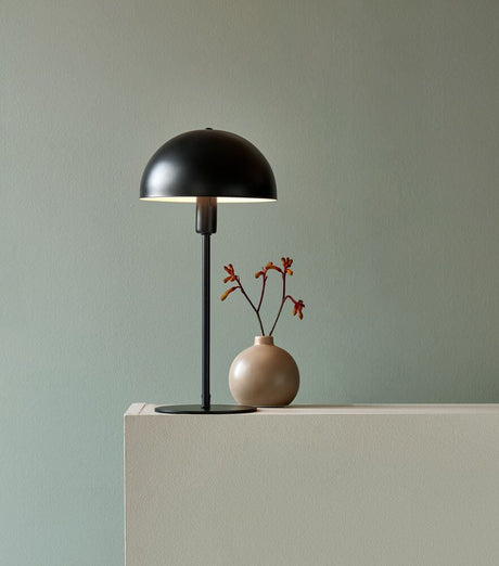 Lampe de table Ellen Noir sur une table d’appoint avec vase et fleurs dans un éclairage doux.