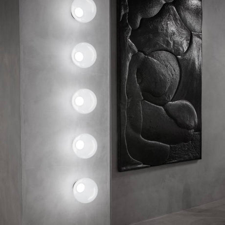 Tom Dixon Opal Wandleuchte Ø 25cm an einer grauen Wand mit modernem Kunstwerk im Hintergrund.