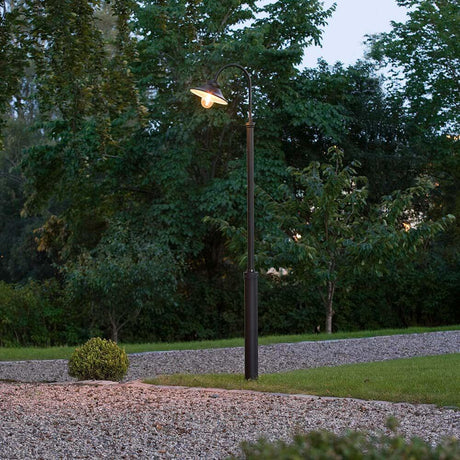 Vega vloerlamp Zwart, helder glas in de tuin bij schemering, omringd door bomen en stenen.