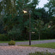 Vega Standleuchte Schwarz, klares Glas im Gartenbereich bei Dämmerung, umgeben von Bäumen und Steinen.