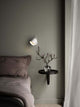 Wandleuchte Angle an einer grauen Wand neben einem Bett mit einem kleinen Tisch und einer blühenden Pflanze.