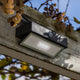 LED-Wandleuchte Arrow mit Solarpanel und Bewegungsmelder an einem Holzbalken montiert, umgeben von Pflanzen.
