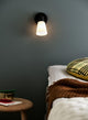 Wandleuchte Cole an der Wand neben einem Bett mit einem gemusterten Kissen und einem Holztisch.
