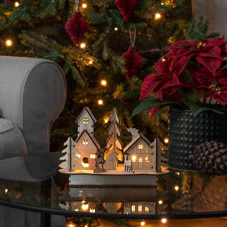 Weihnachtliche Dekoleuchte mit Haus und Tieren aus Holz Braun auf einem Tisch vor einem Weihnachtsbaum.