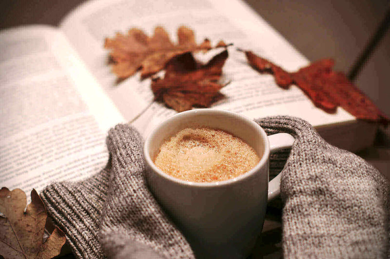 Tasse Kaffee zwischen grauen Handschuhen, umgeben von Herbstblättern und einem geöffneten Buch.