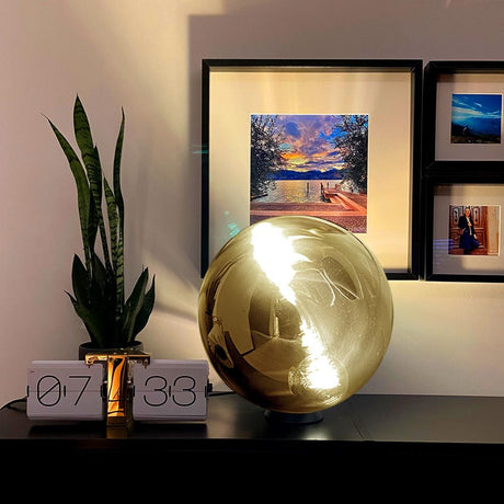 s.luce Progress Glaskugel Tischleuchte auf einem Tisch mit Pflanzen und Kunstwerken im Hintergrund.