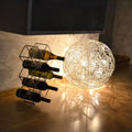 s.luce Mesh Drahtkugel mit warmweißem Licht neben einem Weinregal. Ideal für stimmungsvolle Beleuchtung im Innen- und Außenbereich.