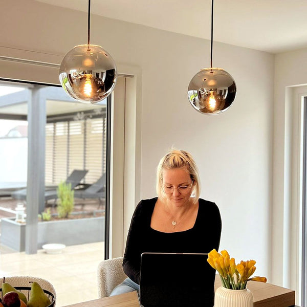 s.luce Orb Lampada a sospensione a sfera di vetro in un ambiente moderno sopra un tavolo con fiori e una donna al laptop.